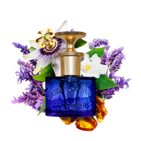 The Caswell Massey Elixir of Love Eau de Toilette features a blue bottle with a gold cap, surrounded by purple flowers, white blossoms, green leaves, and amber stones on a white background. This cruelty-free floral scent exudes elegance.