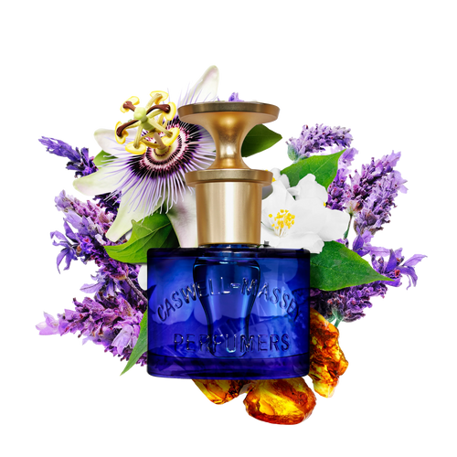 The Caswell Massey Elixir of Love Eau de Toilette features a blue bottle with a gold cap, surrounded by purple flowers, white blossoms, green leaves, and amber stones on a white background. This cruelty-free floral scent exudes elegance.