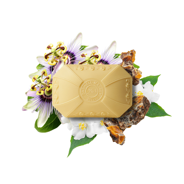 A beige embossed bar from the Caswell-Massey Elixir of Love Three-Soap Set sits on a white background, surrounded by passionflowers, green leaves, and amber stones—bringing plant-based luxury to your skincare routine.