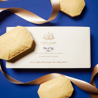 A white Caswell Massey Elixir of Love Three-Soap Set box with gold lettering rests on blue, surrounded by three pale yellow embossed soaps and a gold ribbon. These plant-based triple-milled soaps provide a moisturizing, scented experience.