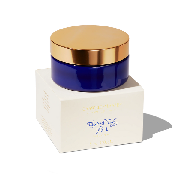 A blue glass jar with a gold lid, labeled “Caswell-Massey Elixir of Love Body Cream,” sits on a white box. This luxurious moisturizing cream by Caswell-Massey features a passionflower fragrance, shown against a plain white background.