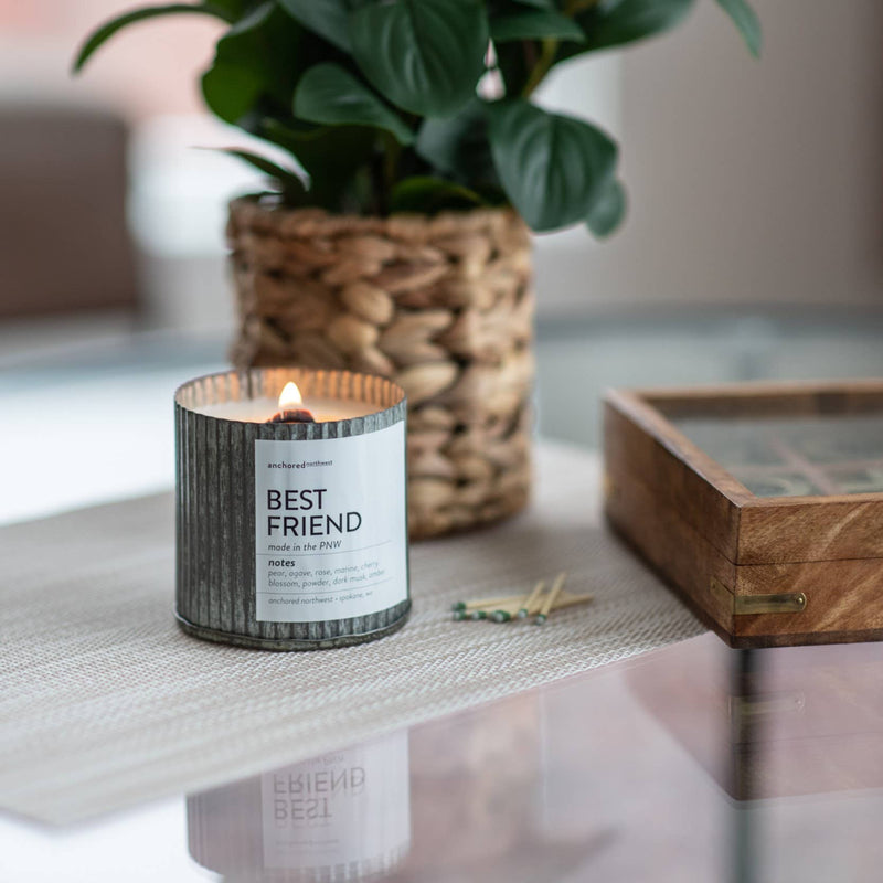 An Anchored Northwest Best Friend candle, housed in a rustic vintage farmhouse container and featuring a wood wick, is lit on a glass table beside a small potted plant and a wooden box. Crafted from American-grown soy wax by Anchored Northwest, this candle creates a warm and cozy ambiance.