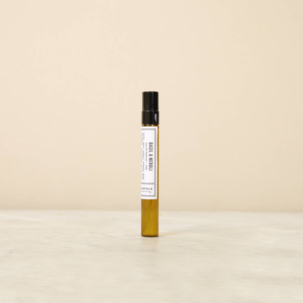 A 10ml amber glass spray bottle of Norfolk Natural Living Basil & Neroli Parfum stands upright on a light marble surface against a beige background, suggesting fresh basil and neroli notes.