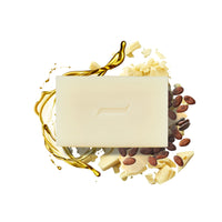 A Caswell-Massey Castile Soap Bar, embossed with a leaf design, is set against a backdrop featuring golden olive oil splashes, cocoa beans, and white chocolate shavings on a white surface.