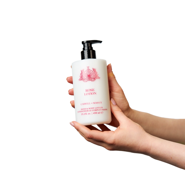 A person with light skin holds a white bottle of Caswell-Massey Rose Hand & Body Lotion with a black pump and pink label against a plain white background.