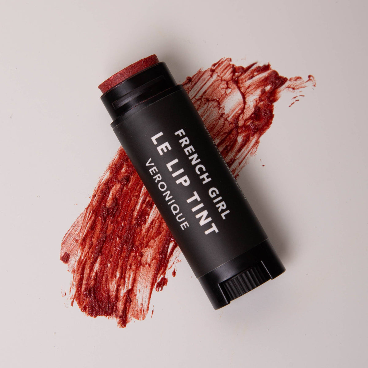 A black tube of FRENCH GIRL Tinted Lip Balm in "Veronique" rests on a white surface, with a swipe of shimmering red balm beneath it, embodying the clean beauty ethos of FRENCH GIRL.