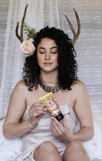 A woman with curly hair sits peacefully, holding a decorated tube of Barefoot Venus Lemon Freckle Shea Butter Lotion. Adorned with a floral arrangement and antlers, she wears a strapless dress and statement necklace, while the air is filled with the fragrance of lemons. A sheer curtain serves as the elegant backdrop.