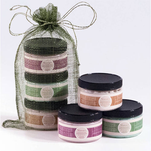 A bundle of jars from Victoria's Lavender 3 Sugar Scrub Set, featuring exfoliating and hydrating sugar scrubs, snugly fits inside a green mesh bag next to three individual jars. The product's vibrant labels in purple, orange, and green make for a striking display against the white background.