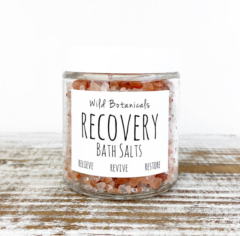 A clear jar labeled "Wild Botanicals Recovery Bath Salts" sits on a wooden surface. The label reads "Relieve, Revive, Restore," hinting at the soothing relief for muscle aches and the infusion of essential oils. The jar is filled with a mix of pink and white bath salts against a white background.