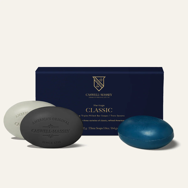 Three oval soaps from the Caswell-Massey Classic Collection Soap Set (white, black, blue) are arranged in front of a dark blue "Heritage Classic" box. These triple-milled, plant-based bars feature gold accents and offer lasting fragrance with refined branding.