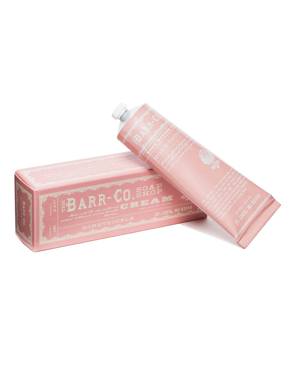 A pink tube of Barr-Co. Honeysuckle Hand & Body Cream, enriched with shea butter and aloe leaf juice, sits partially on its matching pink box—both adorned with white vintage-style text and design elements.