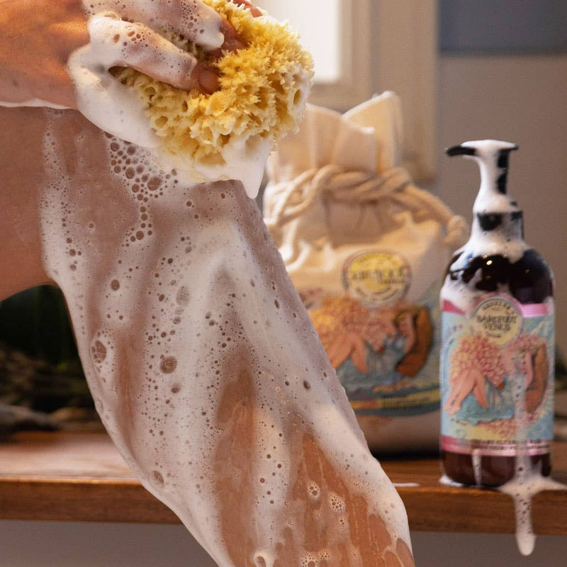 A person scrubs their leg with a yellow sponge, creating a lot of lather from the Barefoot Venus Coconut Kiss Cleansing Hand & Body Wash. In the background, bottles and containers of the all-natural plant-derived formula by Barefoot Venus are placed on a wooden surface near a window. The labels feature colorful designs with flowers.