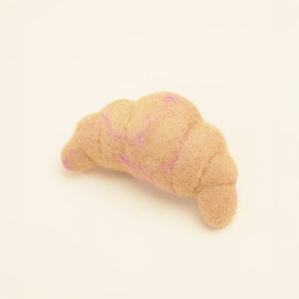 The MODERNBEAST WOOL CROISSANT Wool Dog Toy, a soft, light brown handmade toy with pink accents, is displayed on a plain off-white background.