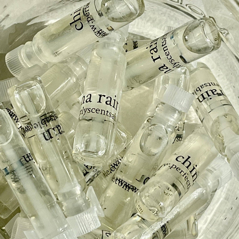 A close-up view of several small, clear glass vials, each filled with a transparent liquid. The vials are labeled "Perfectly Scentsable Perfume Oil Samples" with other smaller text partially visible. These perfume oil vials from the Perfectly Scentsable brand appear to be tightly packed together in a container.