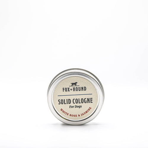 A small circular tin labeled "Fox + Hound White Rose and Jasmine Solid Cologne" is centered on a plain white background. The solid cologne's label features the scent description "White Rose + Jasmine." The tin has a metallic finish and boasts a simple, clean design.