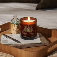 A lit Caswell-Massey Candle - Cognac & Cashmere in a brown glass jar sits on a wooden tray with matches, a pen, and envelopes on a bed with white bedding and brown pillows in the background.