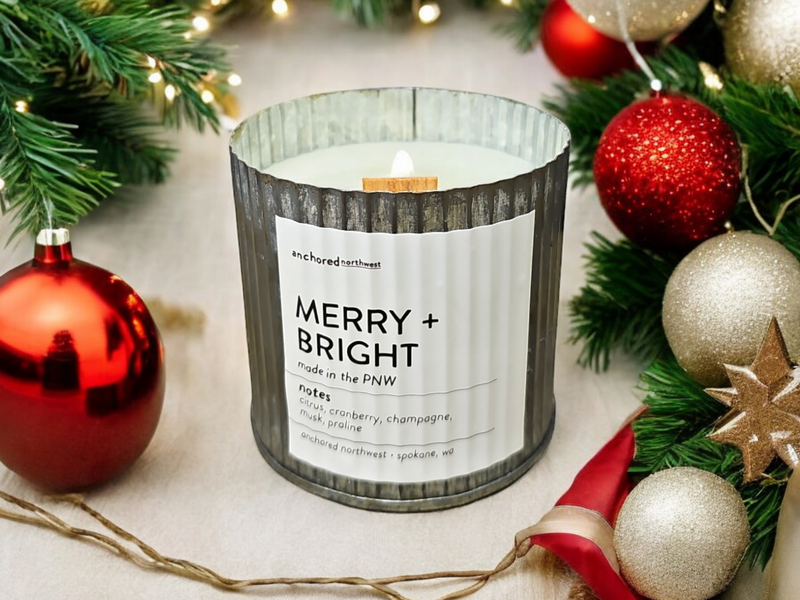 The Anchored Northwest Merry + Bright Rustic Vintage Farmhouse Wood Wick Candle captures rustic charm as it sits amidst evergreen branches and red and gold ornaments. Its soy wax, housed in a ribbed container, releases cranberry and champagne scents, while the wood wick provides a cozy crackle.