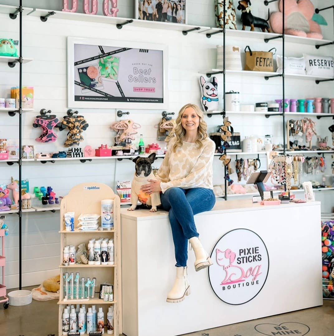 A woman sits on a counter in a pet boutique, surrounded by dog toys and accessories crafted from eco-friendly wheat straw bioplastic. She smiles while holding a small black and white dog, showcasing the significance of pet dental health with the Wag & Bright Supply Co. - Puppy Polisher Eco Toothbrush (Medium). The store's clean, modern aesthetic features colorful products neatly displayed on shelves.