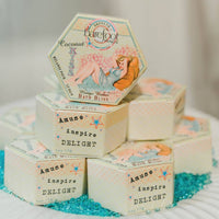 Hexagonal bath product boxes displayed in a pyramid arrangement. The top box is labeled "Barefoot Venus" and features a vintage-style illustration of a woman. Other boxes are labeled "Amuse, inspire, DELIGHT" and highlight natural ingredients like the Barefoot Venus Coconut Kiss Bath Bliss Soak. The background has scattered blue pebbles.