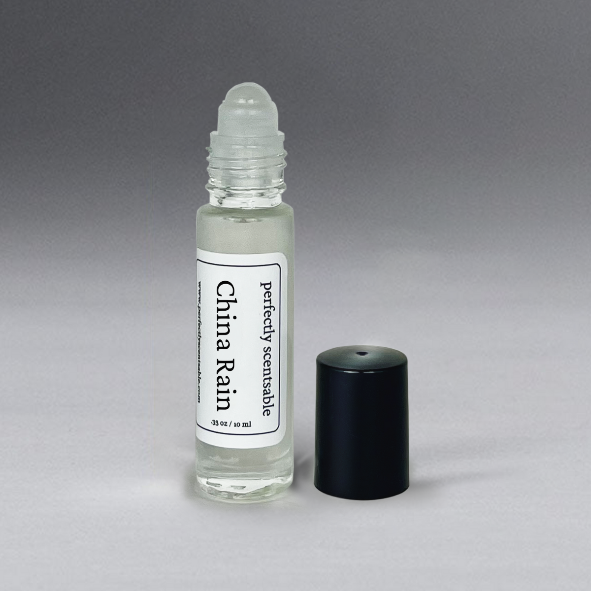 A small clear glass bottle with a rollerball applicator, labeled "Perfectly Scentsable China Rain Perfume Oil - Rollerball." The bottle contains 0.35 fluid ounces (10 milliliters) of liquid, offering an alluring aroma. An uncapped black plastic lid is positioned next to the Perfectly Scentsable China Rain scent on a grey gradient background.