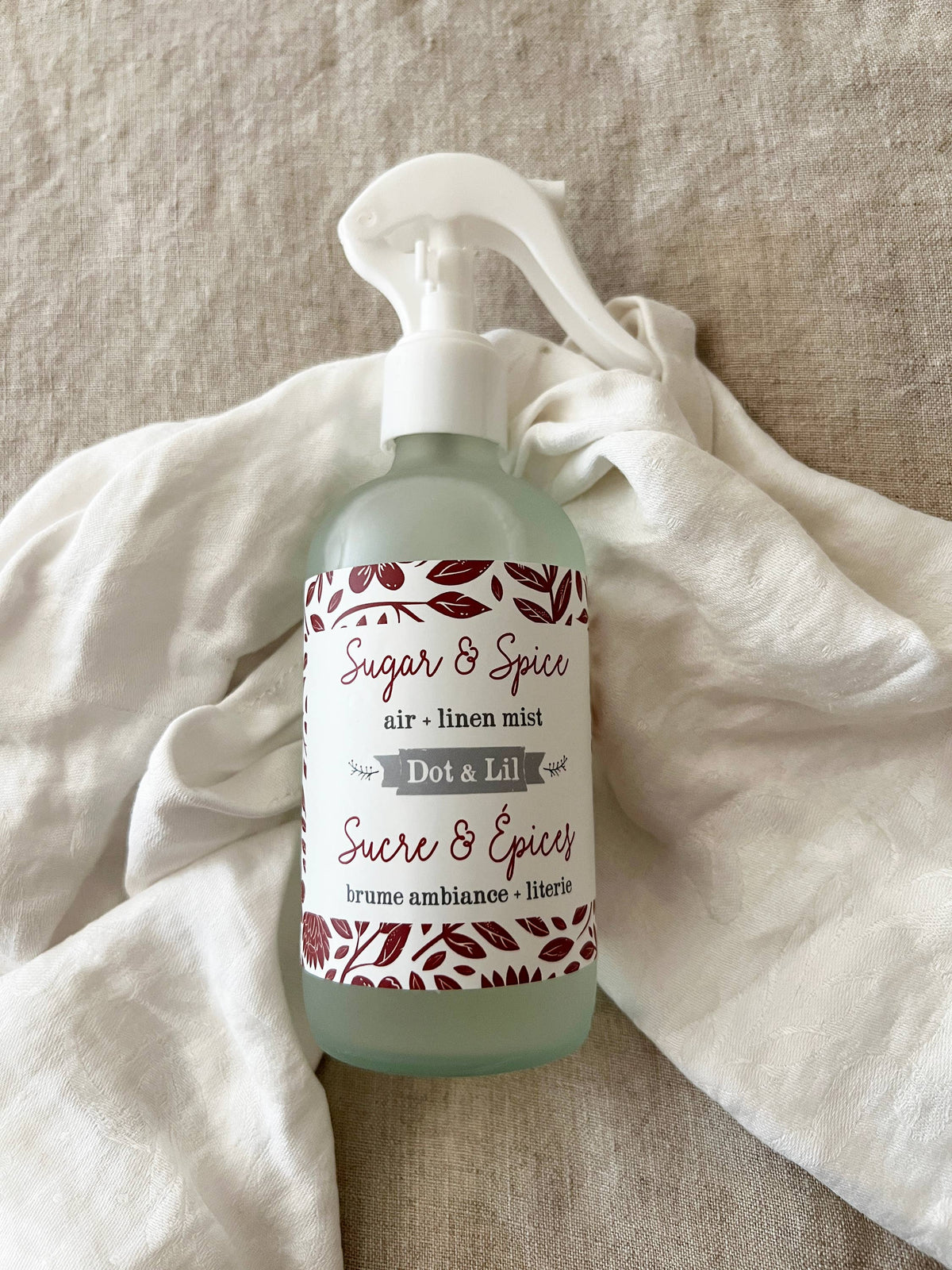A bottle of Dot & Lil Sugar & Spices Linen + Air Mist by Dot & Lil with a spray nozzle sits on a light fabric. The label displays decorative red accents and includes both English and French text.