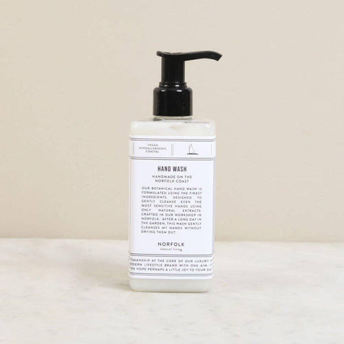 A clear plastic pump bottle of Norfolk Natural Living En Plein Air Natural Hand Wash (300ml) with a white label and black text stands on a light beige surface against a plain beige background. The label details product info and ingredients.