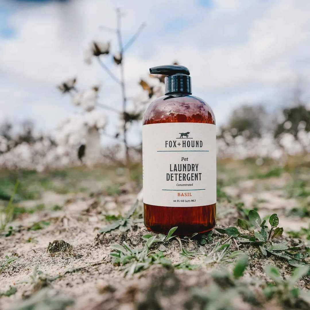 A bottle of Fox + Hound Pet Natural Laundry Detergent Basil with a pump rests outside on sandy ground with green plants, framed by blurred cotton plants and a partly cloudy sky.