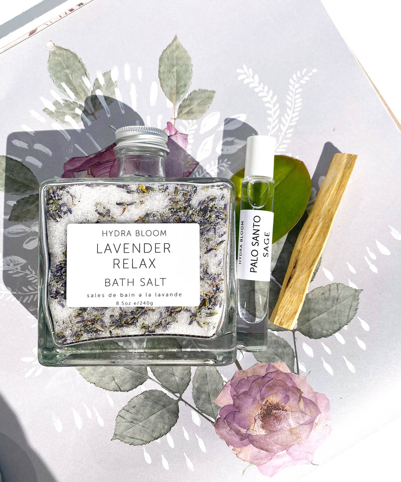 A bottle of Hydra Bloom Beauty Lavender Relax Bath Salts with Epsom & Dead Sea salts, surrounded by dried flowers, a roller oil bottle labeled palo santo, and a piece of wood.