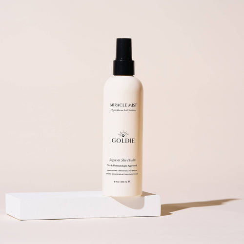 A white spray bottle labeled "Goldie - Miracle Mist" by Goldie stands upright on a white platform against a beige background, with soft side shadows. The veterinarian-approved formula features hypochlorous acid.
