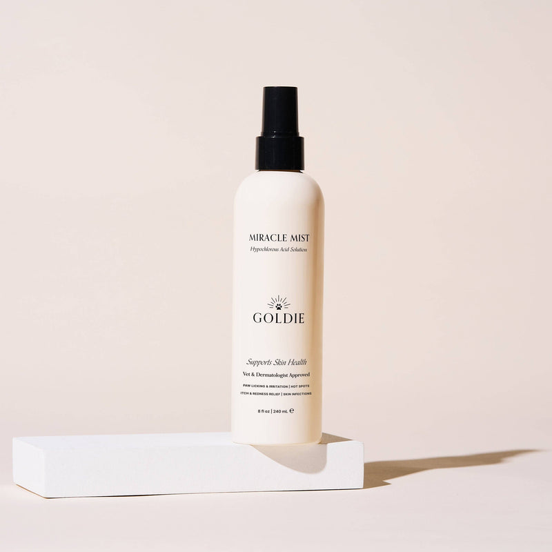 A white spray bottle labeled "Goldie - Miracle Mist" by Goldie stands upright on a white platform against a beige background, with soft side shadows. The veterinarian-approved formula features hypochlorous acid.