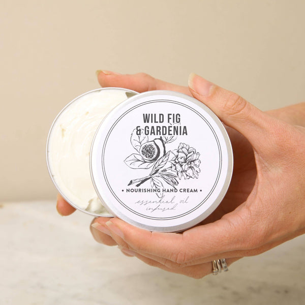 A person holds an open tin of Norfolk Natural Living Wild Fig & Gardenia Gardener's Hand Cream 100ml. The vegan-friendly tin features a floral lid illustration and reveals the nourishing cream inside.