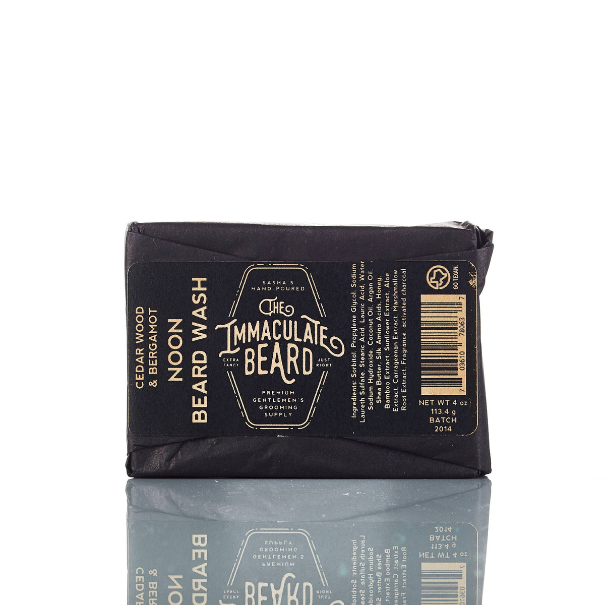 The Immaculate Beard SUNRISE Beard Wash Bar, wrapped in black paper with gold and white text listing the product name, ingredients, and brand, rests on a white surface with its reflection visible below.