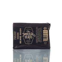The Immaculate Beard SUNRISE Beard Wash Bar, wrapped in black paper with gold and white text listing the product name, ingredients, and brand, rests on a white surface with its reflection visible below.
