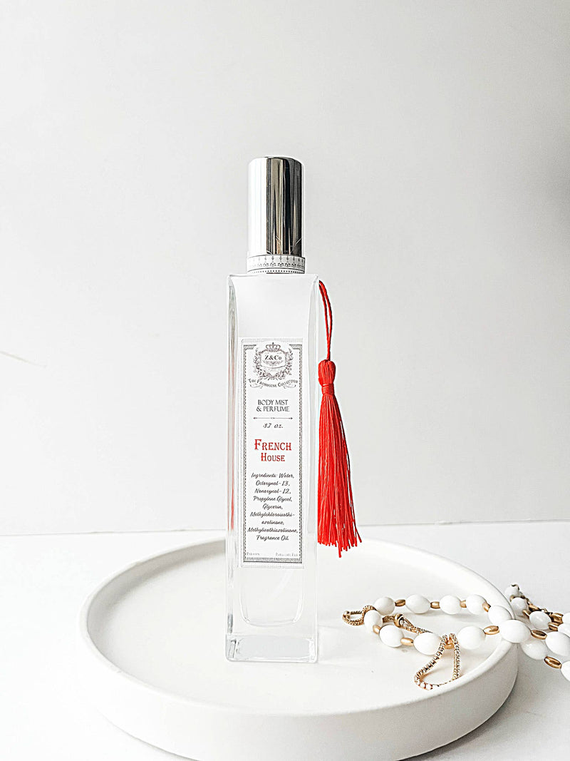 The Z&Co. Farmhouse Collection Body Mist Perfume, in a clear glass bottle with a silver cap and red tassel, rests on a white tray beside a gold-accented beaded bracelet—ready to add lasting fragrance to your day.