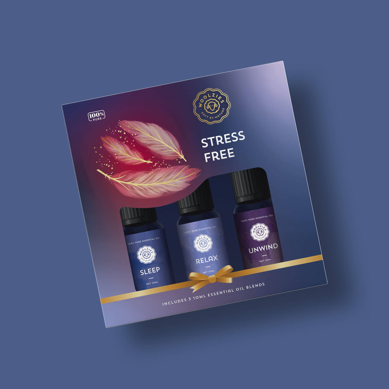 The Woolzies Stress Free Holiday Collection by Woolzies is a gift box featuring three essential oil blends—SLEEP, RELAX, and UNWIND—on a blue background with "STRESS FREE," feather illustrations, and a gold ribbon graphic. Perfect for relaxation.