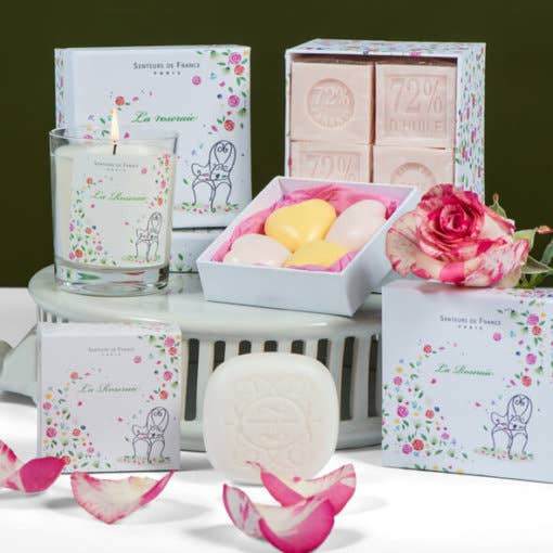 A collection of Senteurs De France bath and body products including "Roseraie" Honeysuckle & Rose heart soaps, a candle, and bath bombs, displayed with a rose and petals, all set against a green background.