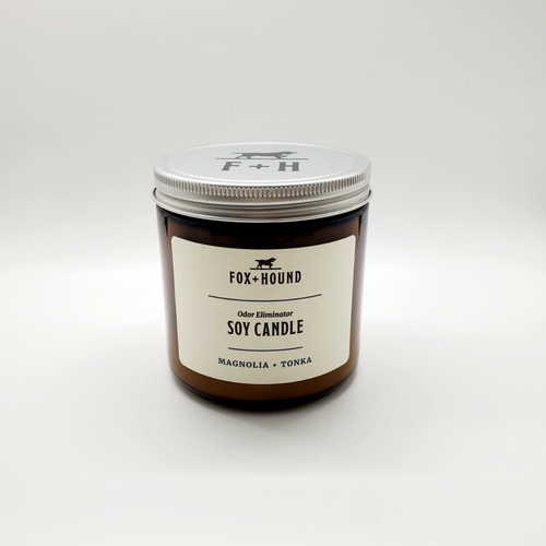 A brown glass jar candle with a silver lid labeled "Fox + Hound - Odor Eliminator Soy Candle - Magnolia + Tonka" effectively tackles unwanted odors, filling the air with the delightful scent of Magnolia and Tonka against a white background.