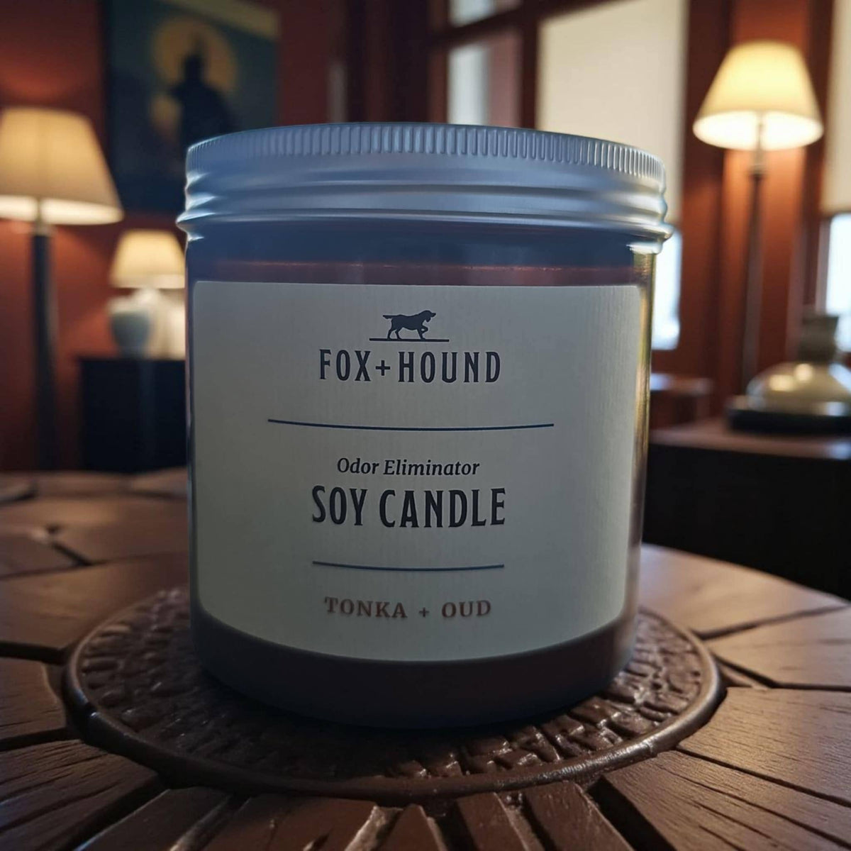 A Fox + Hound Odor Eliminator Soy Candle - Tonka & Oud rests on a wooden table in a cozy room, blending luxurious scents and effective pet odor removal from the trusted Fox + Hound brand.