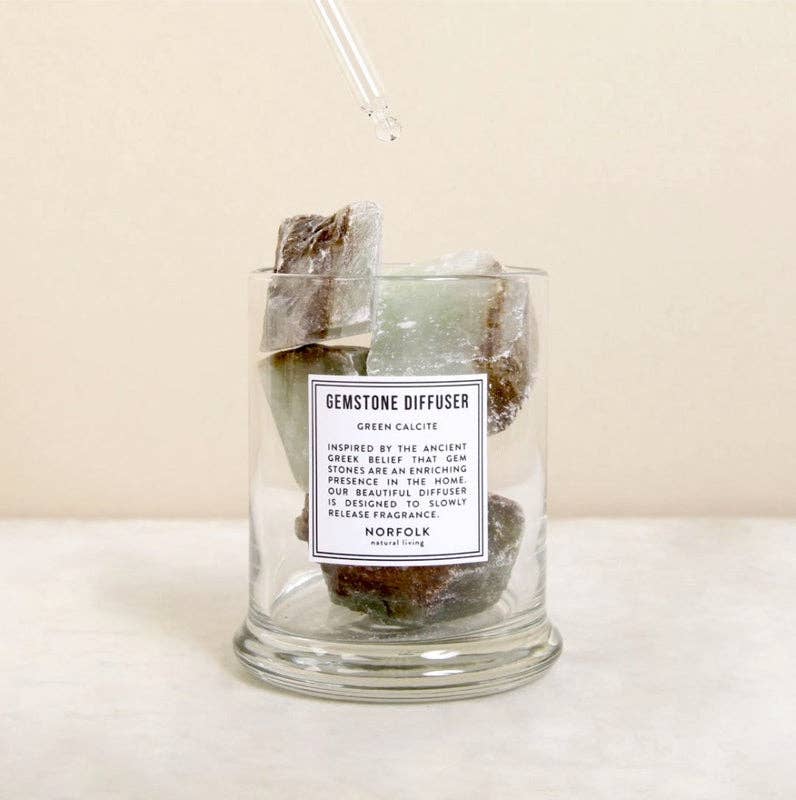 A Norfolk Natural Living Gemstone Green Calcit Diffuser glass jar sits on a light surface with large green and brown stones inside. A dropper releases oil onto the stones, set against a neutral background.