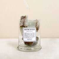 A Norfolk Natural Living Gemstone Green Calcit Diffuser glass jar sits on a light surface with large green and brown stones inside. A dropper releases oil onto the stones, set against a neutral background.