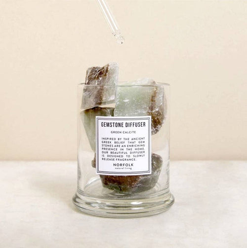 A Norfolk Natural Living Gemstone Green Calcit Diffuser glass jar sits on a light surface with large green and brown stones inside. A dropper releases oil onto the stones, set against a neutral background.