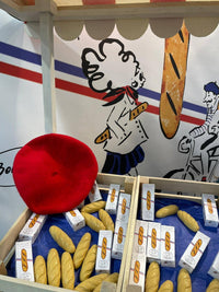 A market stall display features Senteurs De France’s “Baguette” Soap and boxes, a red beret, and cartoon art of a beret-wearing figure with a baguette—ideal as a French keepsake or for charming French-themed decor.