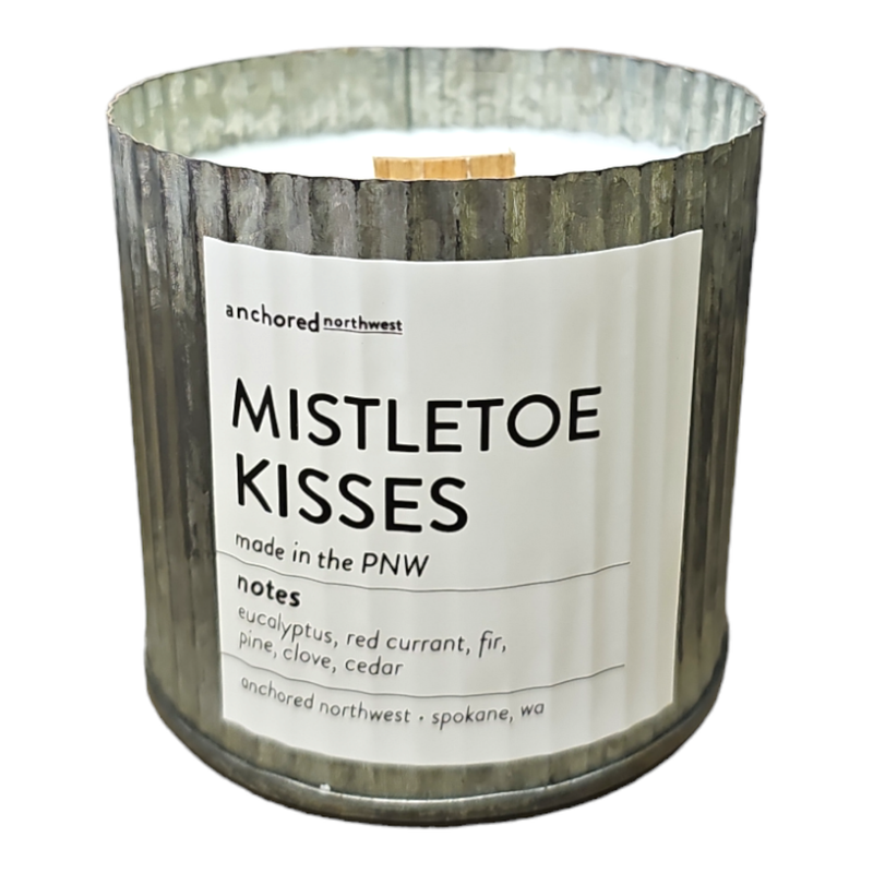 A rustic vintage farmhouse wood wick soy wax candle by Anchored Northwest, presented in a textured metallic holder and labeled "Mistletoe Kisses." The label highlights scents of eucalyptus, red currant, fir, pine, clove, and cedar. Made in the PNW.