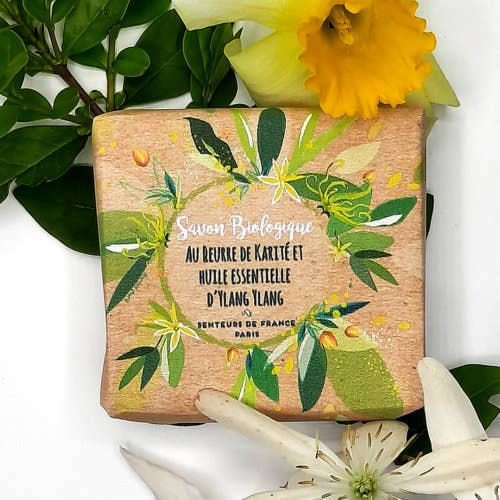 Senteurs De France - Organic soap ylang ylang essential oil by Senteurs De France is wrapped and surrounded by green leaves plus yellow and white flowers, highlighting its organic ingredients and soothing fragrance.