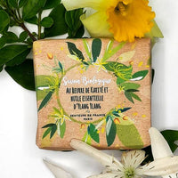 Senteurs De France - Organic soap ylang ylang essential oil by Senteurs De France is wrapped and surrounded by green leaves plus yellow and white flowers, highlighting its organic ingredients and soothing fragrance.