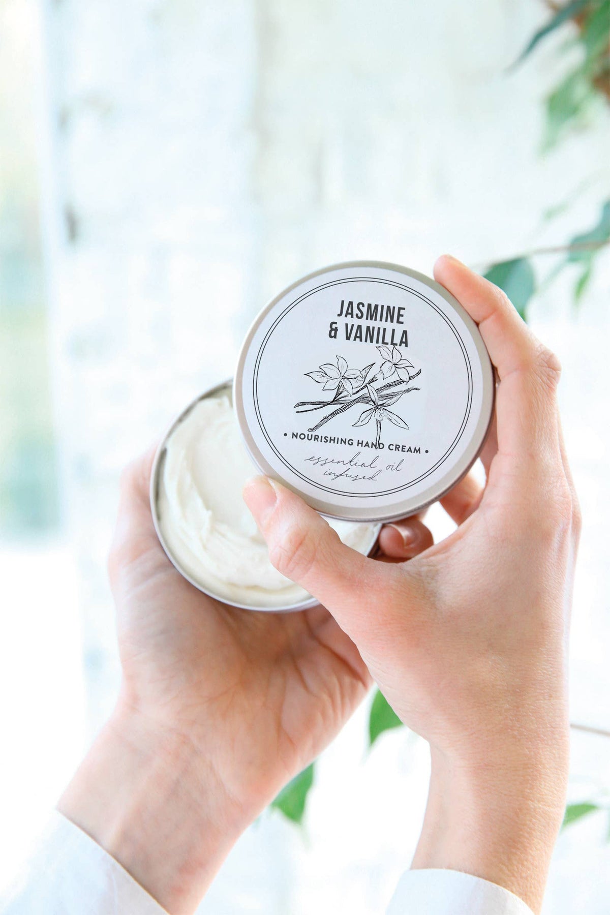 A person holds an open tin of Norfolk Natural Living Jasmine & Vanilla Gardener's Hand Cream (100ml), showing the creamy contents. The lid displays botanical illustrations and text, with green leaves and bright natural light in the background.