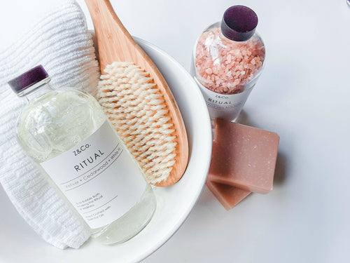 A white bowl contains a bottle of Z&Co. Ritual Market Collection Bubble Bath, a wooden bath brush, a glass jar of pink bath salts, a white towel, and a bar of pink soap. The items are arranged neatly, suggesting a spa or bubble bath setting.