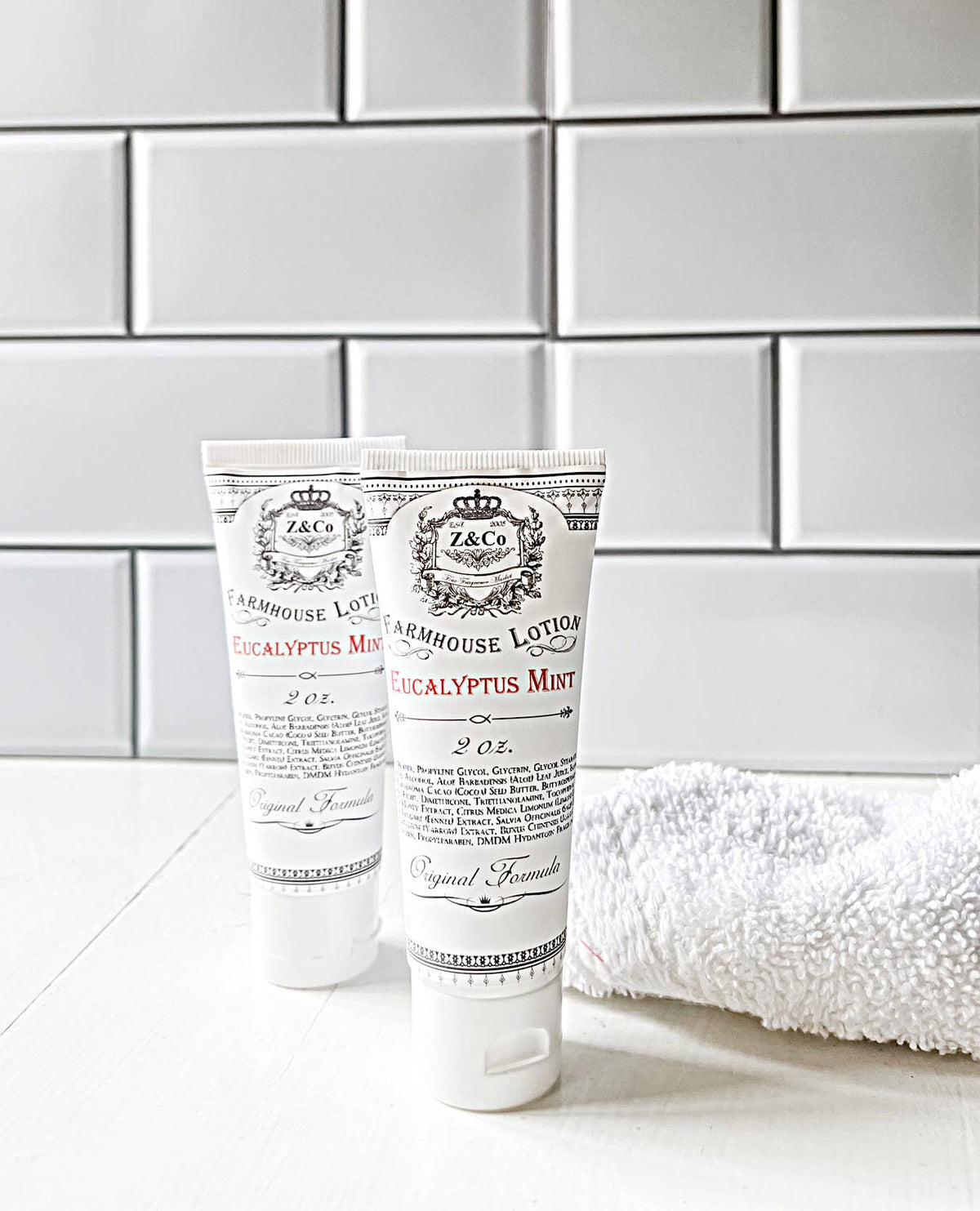 Two tubes of Z&Co. Eucalyptus Mint Farmhouse Lotion Tube 2 oz are placed on a white surface in front of a white subway tile background. The tubes are labeled with product information, and a white towel is positioned to the right.