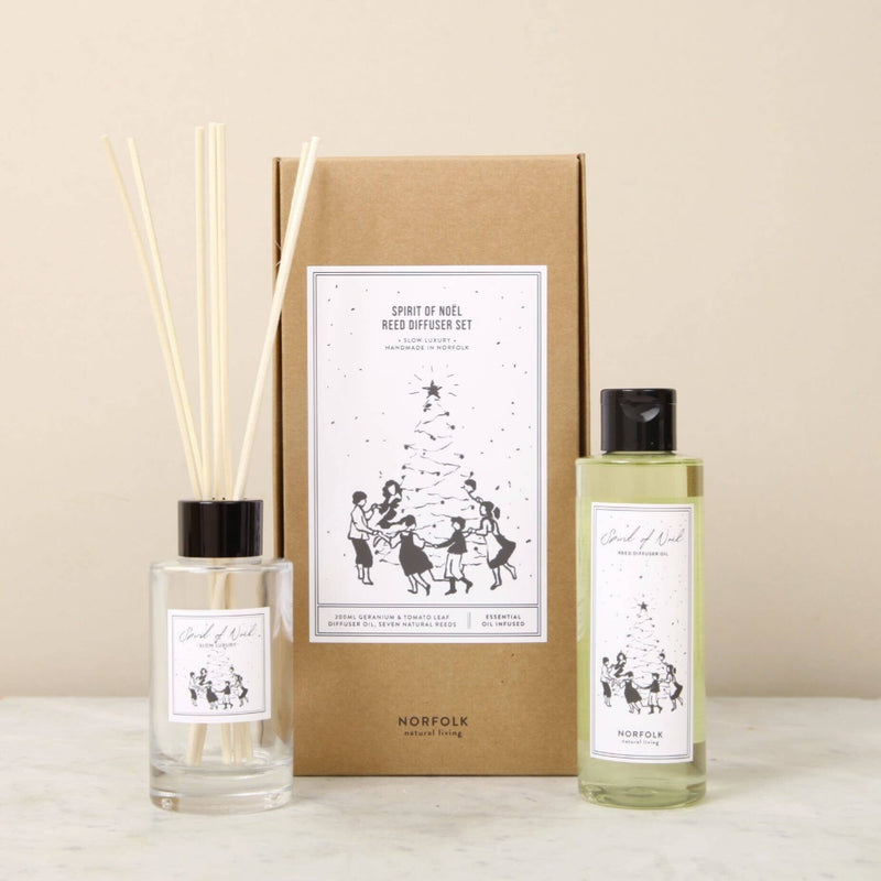The Norfolk Natural Living Spirit Of Noel Reed Diffuser Set includes a glass bottle with reeds, a refill, and a box, all featuring minimalist black and white illustrations of people dancing around a Christmas tree.