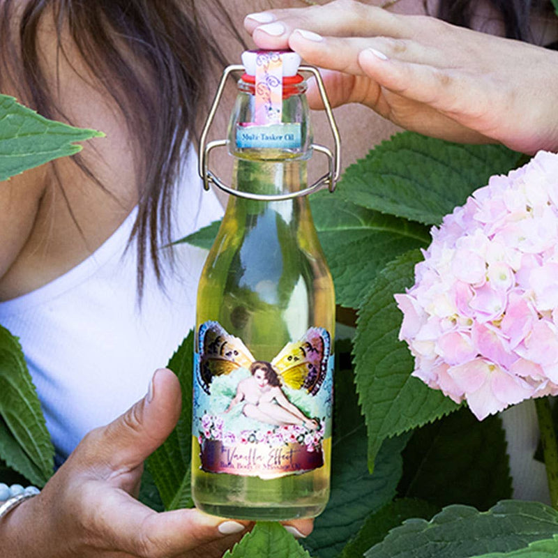 A person holding a bottle of Barefoot Venus The Vanilla Effect Body+Bath+Massage Oil, filled with a yellow liquid infused with natural oils, features a colorful label showcasing a person and butterflies. The scene is framed by green leaves and pink hydrangea flowers.